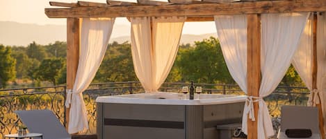 Ein Whirlpool mit einer Pergola und Vorhängen, zwei Liegestühle, ein kleiner Tisch mit einer Pflanze und Blick auf die Landschaft.