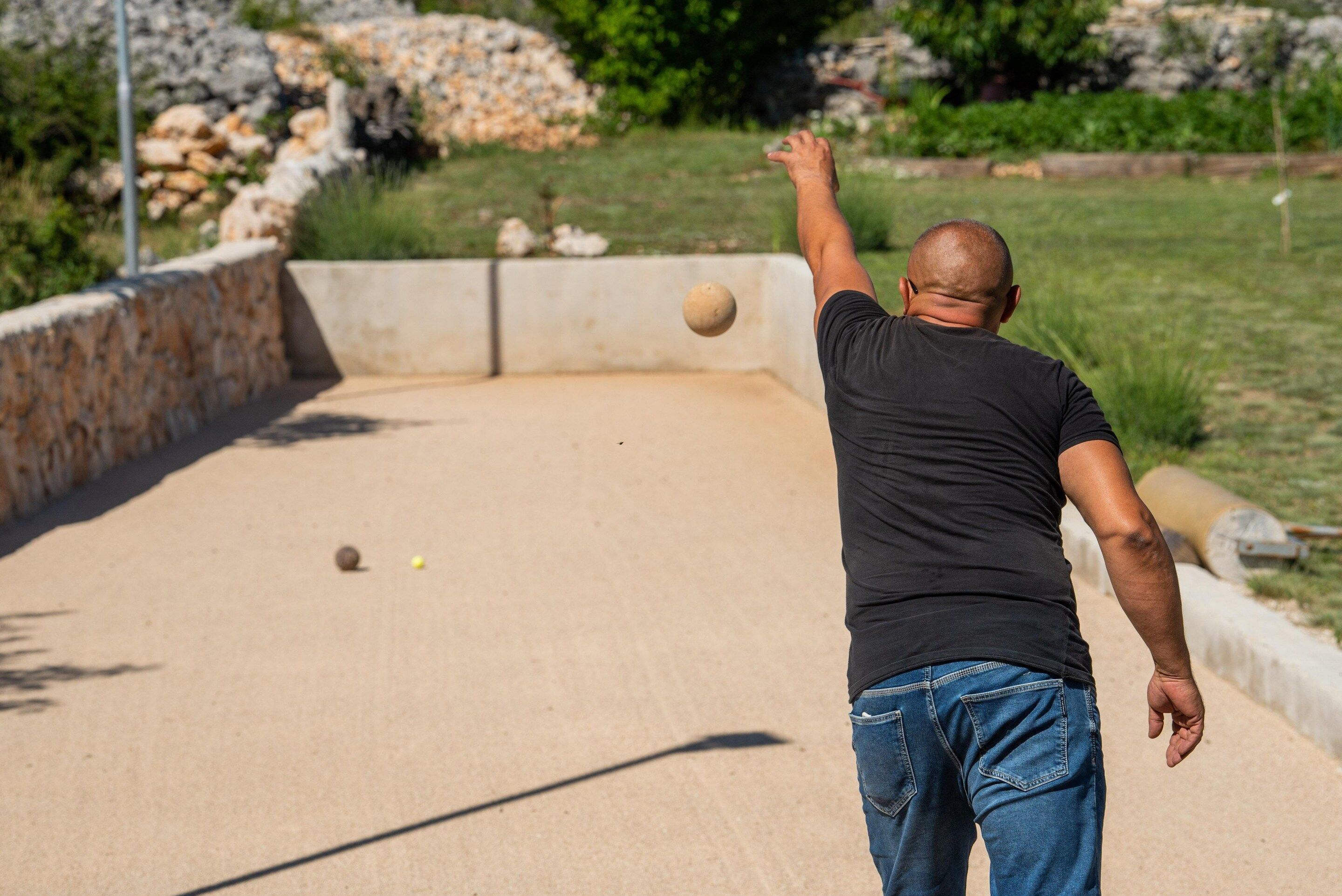 Eine Person spielt Boule auf einer ebenen, offenen Fläche, im Hintergrund eine Steinmauer und Grünpflanzen.