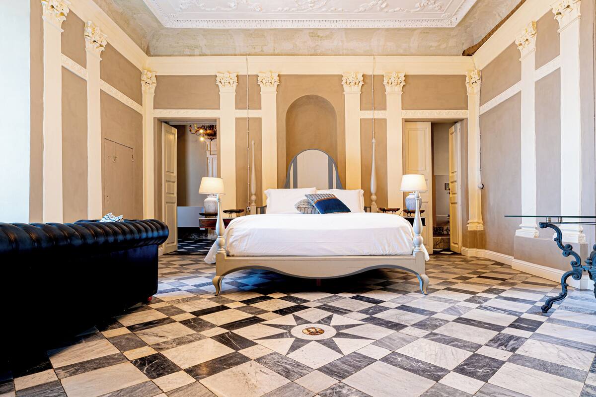 Una habitación de hotel con piso a cuadros, una cama con ropa blanca, un sofá negro y una mesa de vidrio.