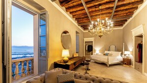 Une chambre avec un grand lit, un coin salon avec un canapé, un lustre et une vue sur la mer.