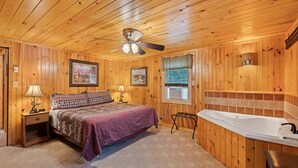 Une chambre dans une cabane en bois, comprenant un lit, une table de chevet, une lampe, un ventilateur de plafond, une fenêtre et une salle de bain avec une baignoire.