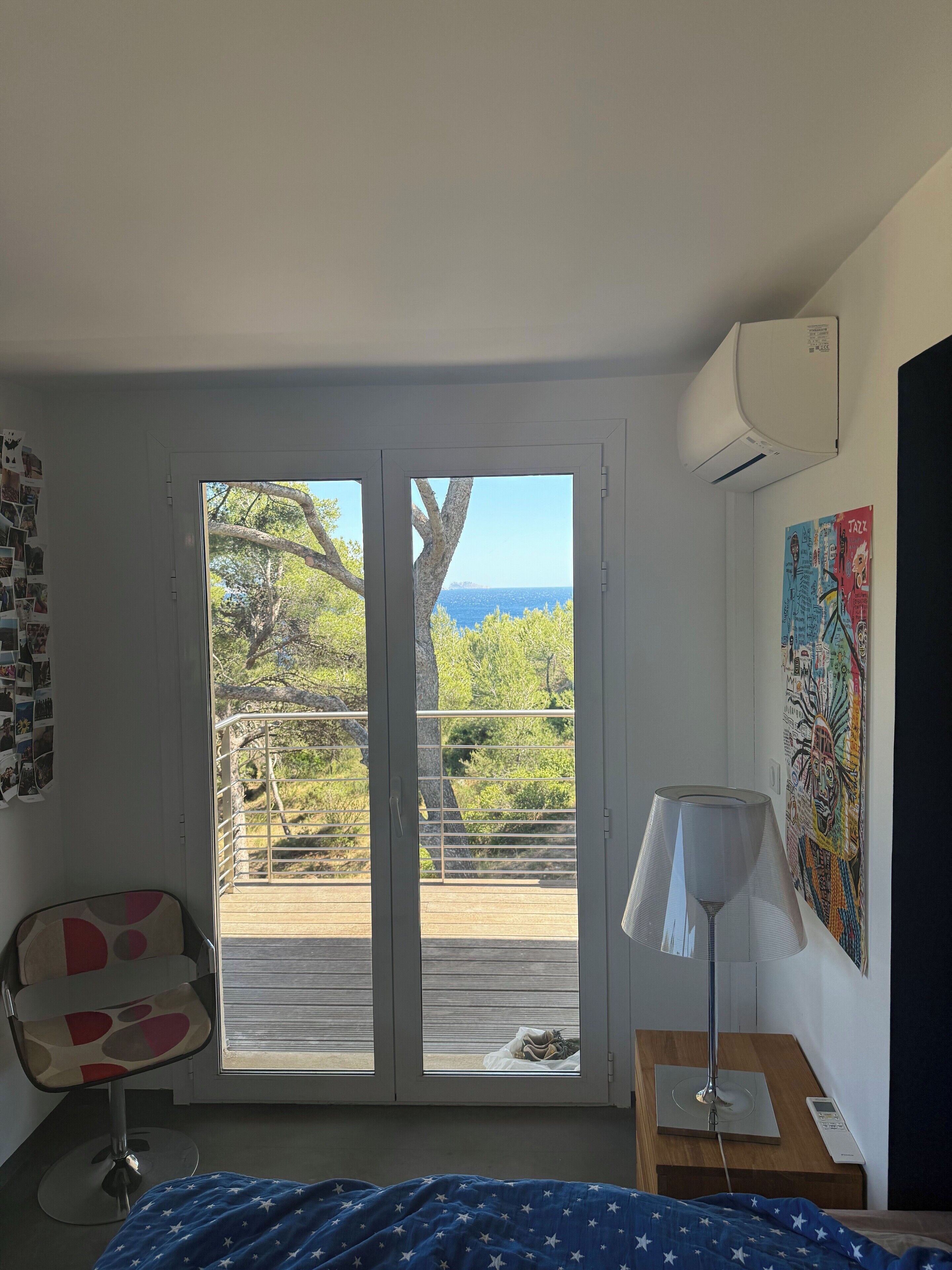 A bedroom with a bed, a chair, a nightstand with a lamp, a wall-mounted air conditioner, and a balcony with a view of trees and the sea.