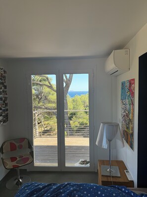 Ein Schlafzimmer mit Bett, einem Sessel, einem Nachttisch mit Lampe, einer Wand-Klimaanlage und einem Balkon mit Blick auf Bäume und das Meer.