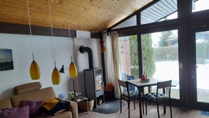 Una sala de estar acogedora con sofá, una mesita de centro de vidrio, una mesa de comedor con sillas, chimenea y ventanas amplias con vistas al exterior.