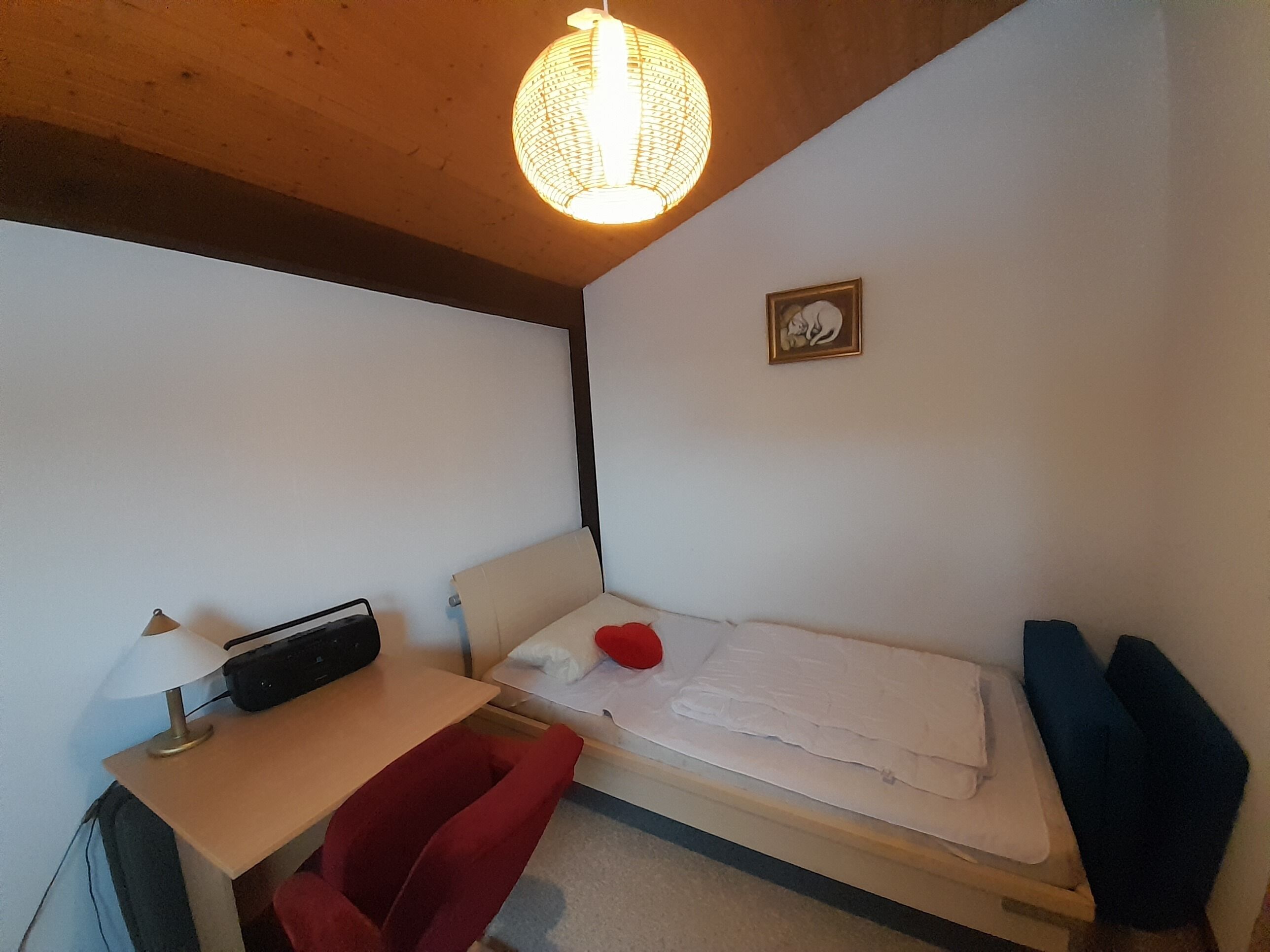 Une petite chambre à coucher, bien rangée, avec un lit simple, un bureau doté d’une lampe et une chaise.