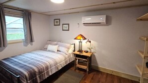 A small bedroom with a bed, a nightstand with a lamp, an air conditioner, and a window with curtains. - Creekside Delight-Lovely! 20 min to Asheville (Arden)