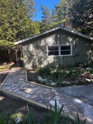 A small, single-story house with a stone pathway leading to the front door, surrounded by a garden with various plants and flowers. - Cozy 1-bedroom cottage in beautiful Fish Creek (Fish Creek)