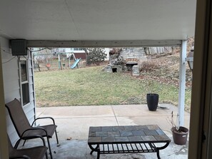 A covered patio with a stone table and two chairs, a potted plant, and a view of a backyard with a playset and stone wall. - Peaceful duplex house in Omaha perfect for a relaxing getaway (Omaha)