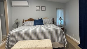 Un dormitorio con cama, mesitas de noche, lámpara, ventana con cortinas y aire acondicionado.