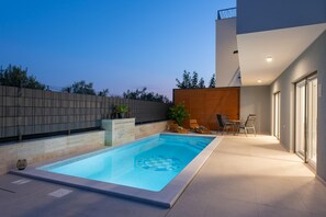 A modern outdoor swimming pool with a checkered pattern, surrounded by a low wall, a wooden deck, and a seating area with chairs and a table. - Holiday Oasis With Hydromassage Pool for 8 Guests (Kastela)