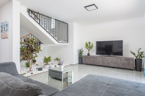 Ein modernes Wohnzimmer mit einem grauen Sofa, einem Glastisch, einem Fernseher auf einer Holzkommode, einer Treppe und Topfpflanzen.