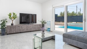 Un soggiorno moderno con televisione a schermo piatto, tavolino in vetro, divano grigio e porta scorrevole in vetro che dà su una piscina.