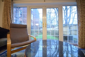 Ein Zimmer mit Glastisch, braunem Stuhl, einer Couch und Blick durch französische Türen in einen Garten.