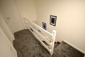 Una escalera con barandilla blanca y suelo alfombrado. Dos cuadros enmarcados adornan la pared.