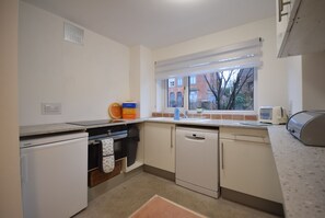 A compact kitchen with a dishwasher, a small refrigerator, a window with a view of a brick building, and a toaster on the counter. - Cosy 2BDR House with Driveway Near Manchester (Manchester)