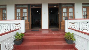 Entrada de prédio com escadaria vermelha, corrimãos brancos e duas plantas em vasos.