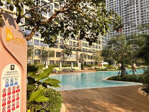 A swimming pool area with lounge chairs, a wooden deck, and a building with balconies in the background. - Vinhomes Grand Park-Masterise Riverside (Ho Chi Minh City)