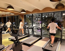 Academia com esteiras, equipamentos de exercício e amplas janelas com vista para a cidade.