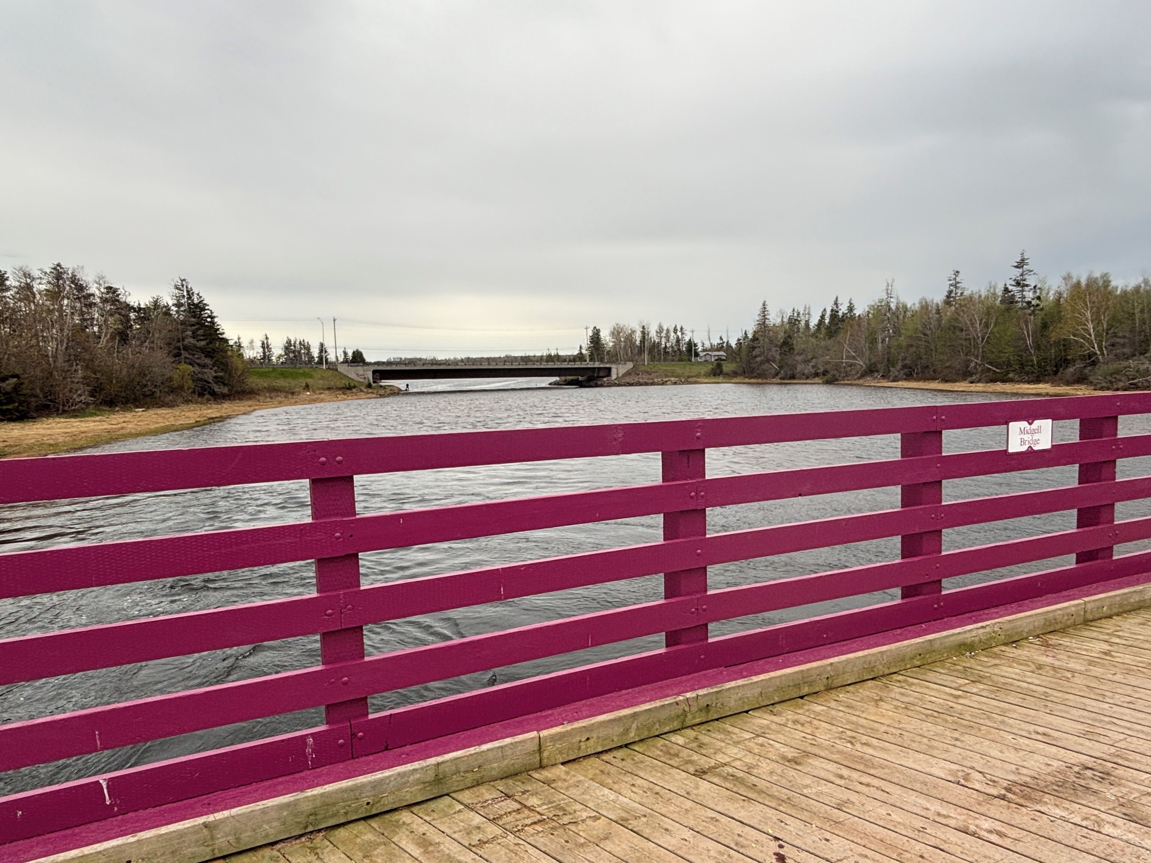 A wooden bridge with a sign reading 'Midgell Bridge' over a river, surrounded by trees and a cloudy sky.