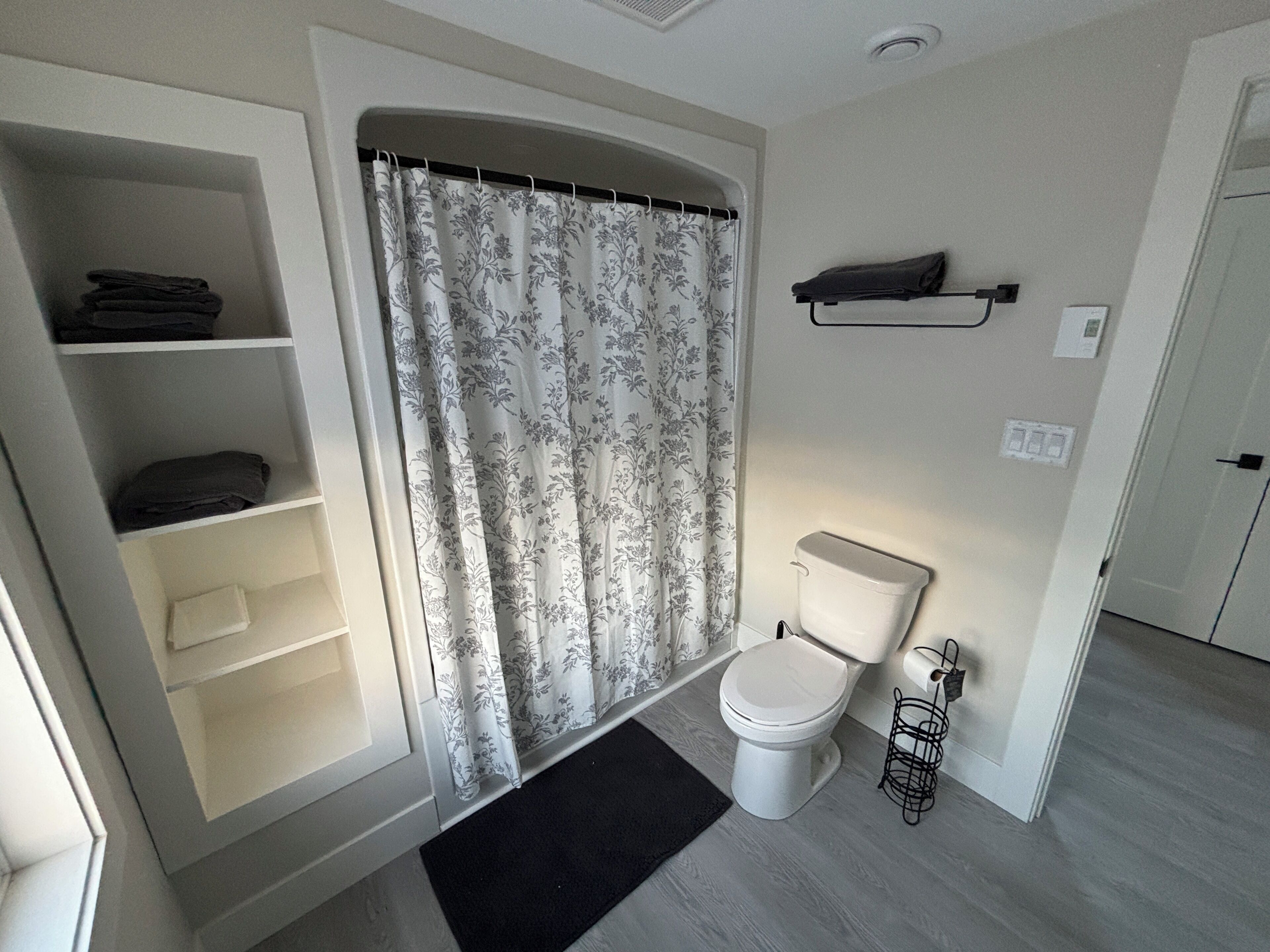 A bathroom with a toilet, a shower curtain, a shelf with folded towels, and a black mat on the floor.