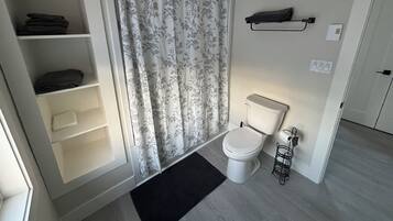 A bathroom with a toilet, a shower curtain, a shelf with folded towels, and a black mat on the floor.