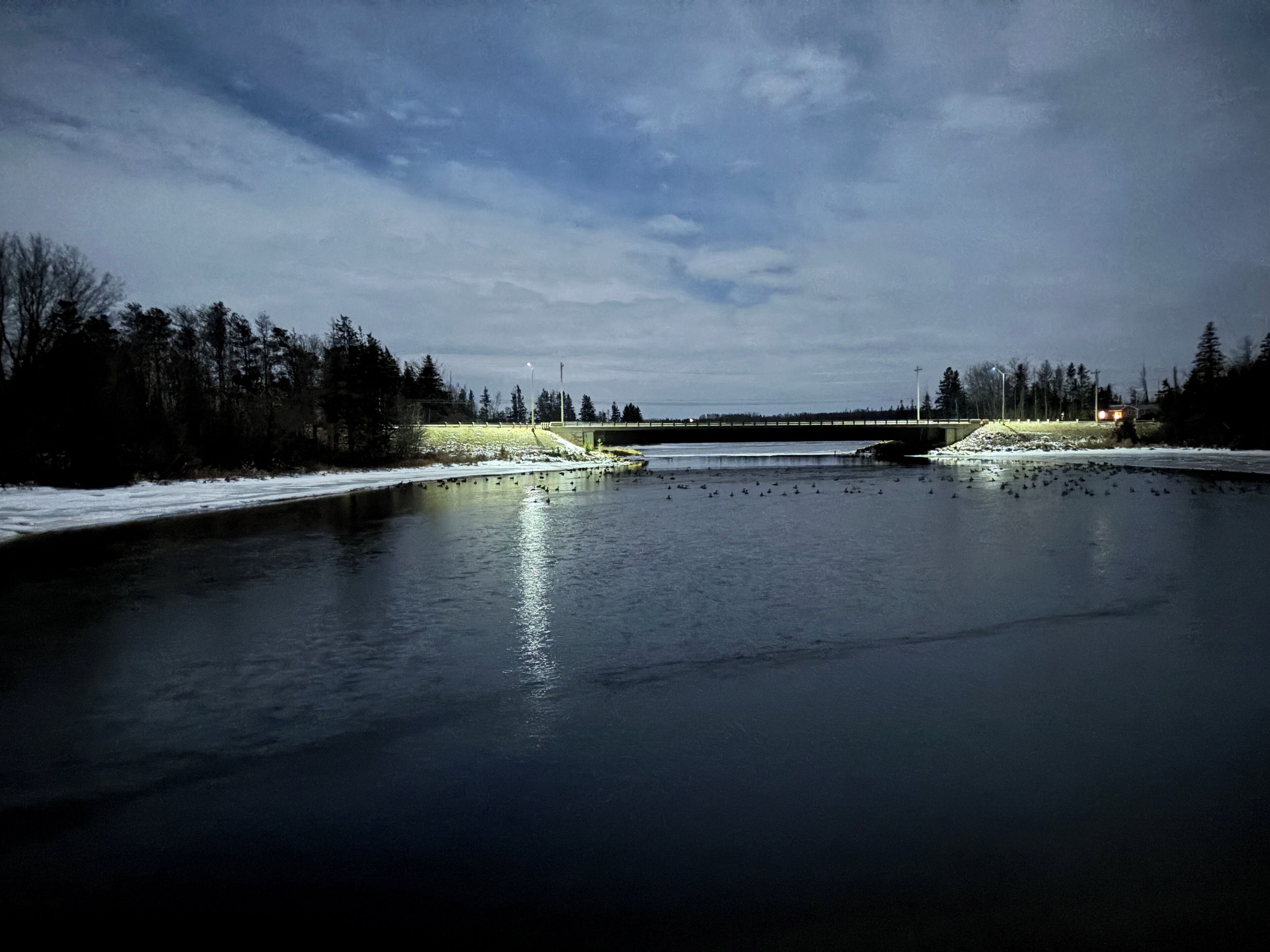 A frozen river with a bridge in the distance, surrounded by trees and snow.
