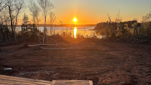 Eine Holzterrasse mit Tisch und zwei Stühlen mit Blick auf ein Gewässer und Bäume bei Sonnenuntergang.