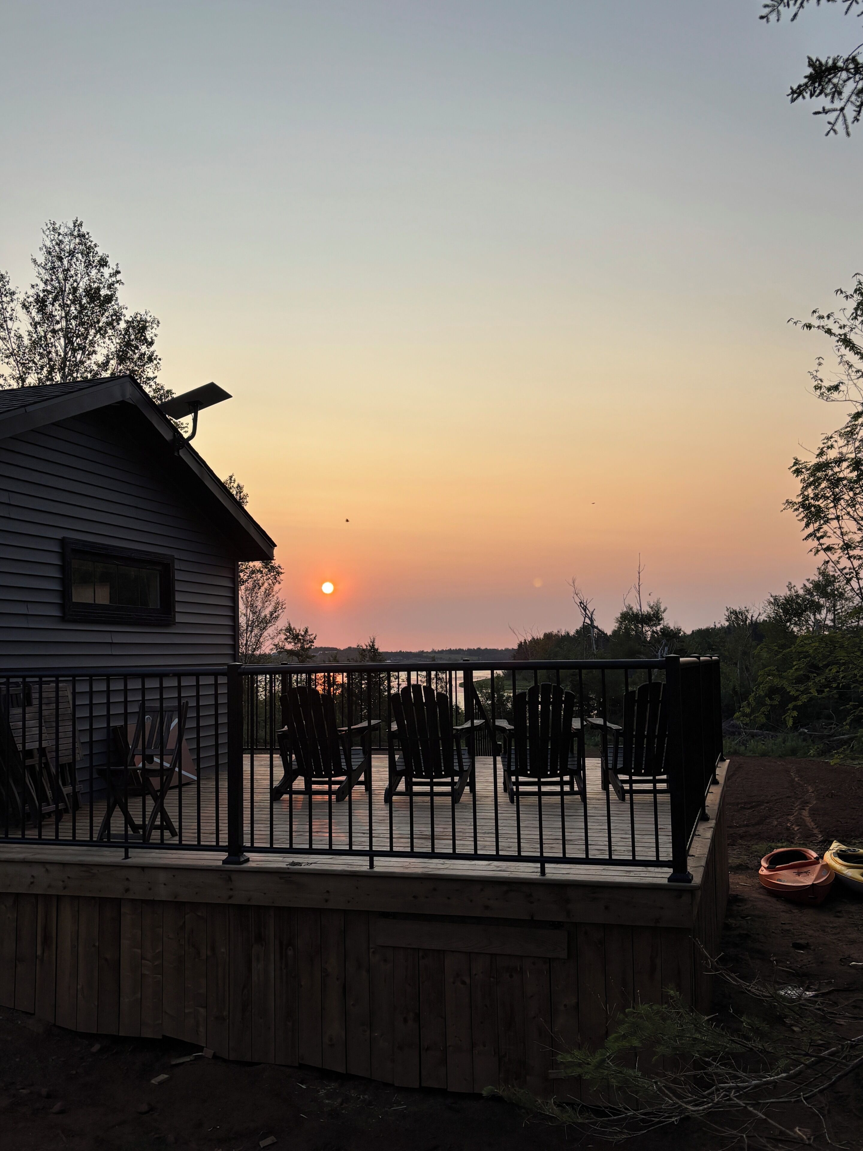 A wooden deck with chairs and a table, a house with a visible garage, and a sunset in the background.
