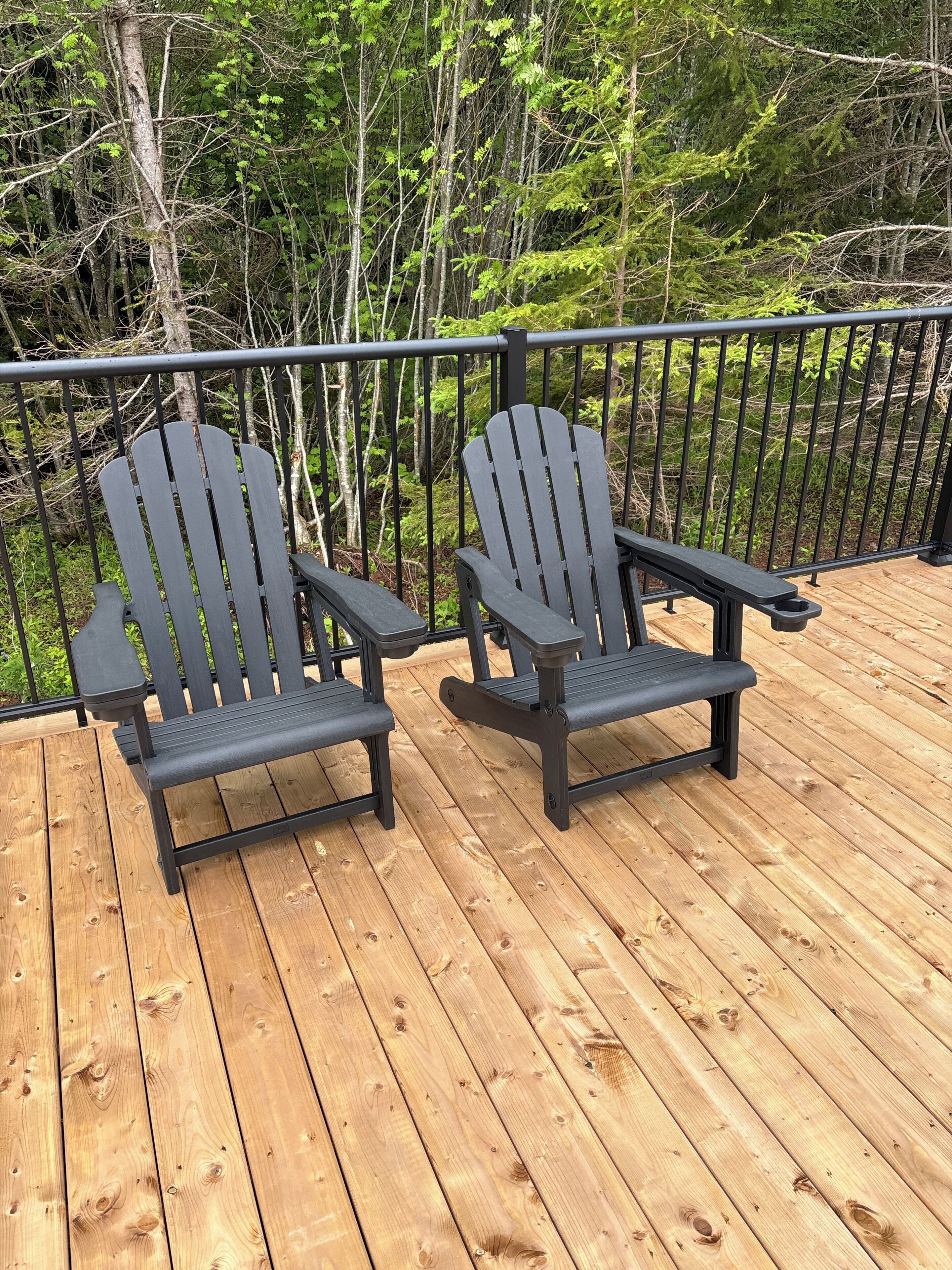 Dos sillas Adirondack en una terraza de madera con baranda de metal, rodeadas de árboles.