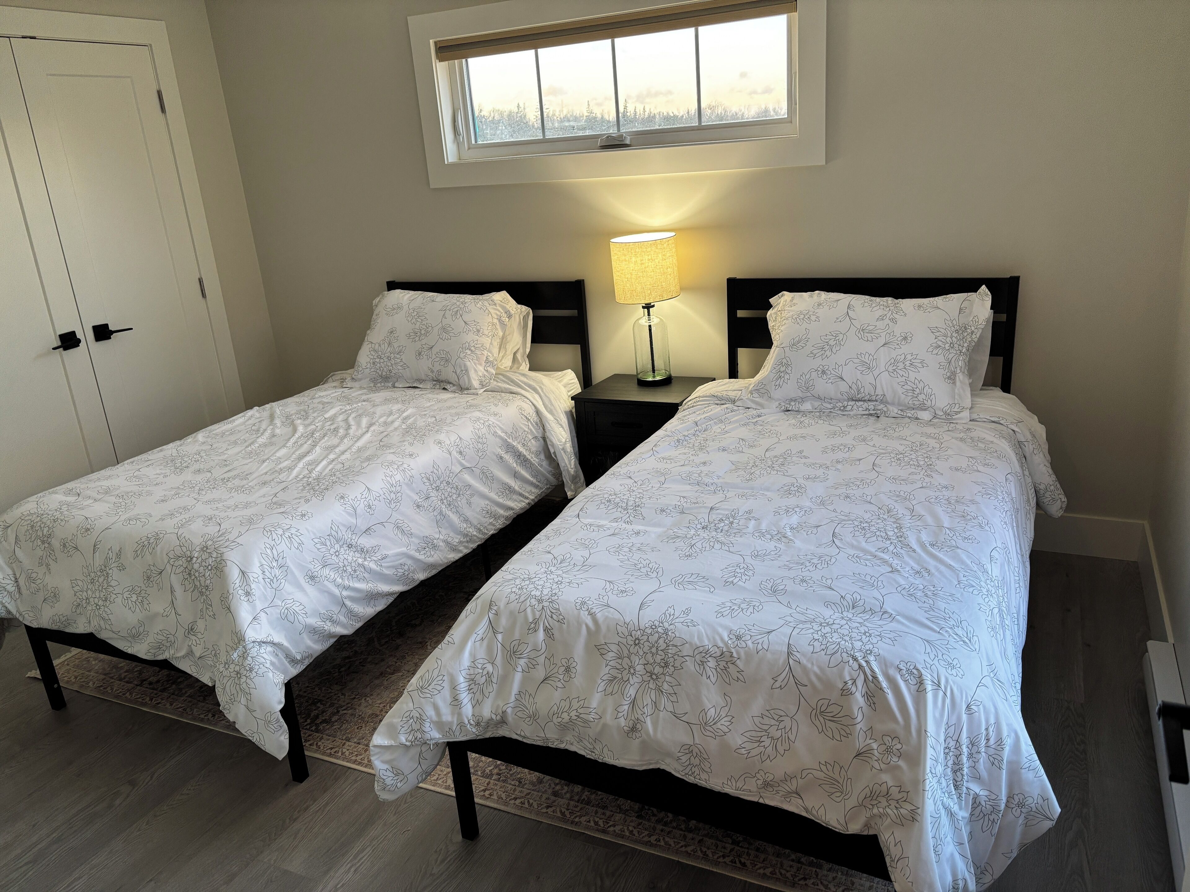 Quarto com duas camas, mesa de cabeceira com luminária, janela com vista para árvores e porta.