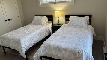 Quarto com duas camas, mesa de cabeceira com luminária, janela com vista para árvores e porta.