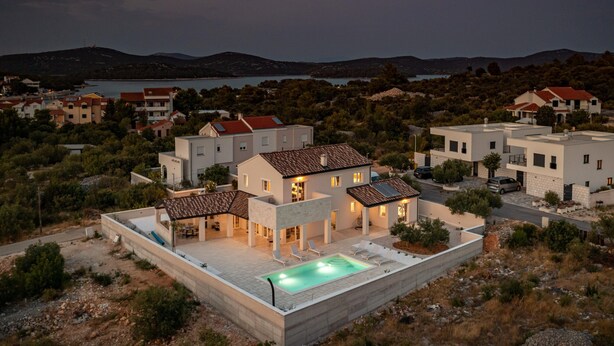 Une villa moderne dotée d’une piscine, entourée d’autres maisons, avec en toile de fond des collines et un lac.