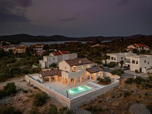 A modern villa with a swimming pool, surrounded by other houses, set against a backdrop of hills and a lake. - Villa Royal Stone With Pool in Jezera (Tisno)