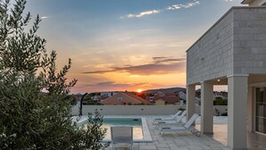 Un espace piscine aménagé sur le toit, avec des chaises longues, une piscine et une vue sur le coucher de soleil au-dessus d’un quartier résidentiel.