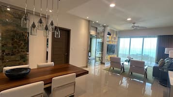 A modern living room with a wooden dining table, white chairs, a sofa, and a TV mounted on the wall.