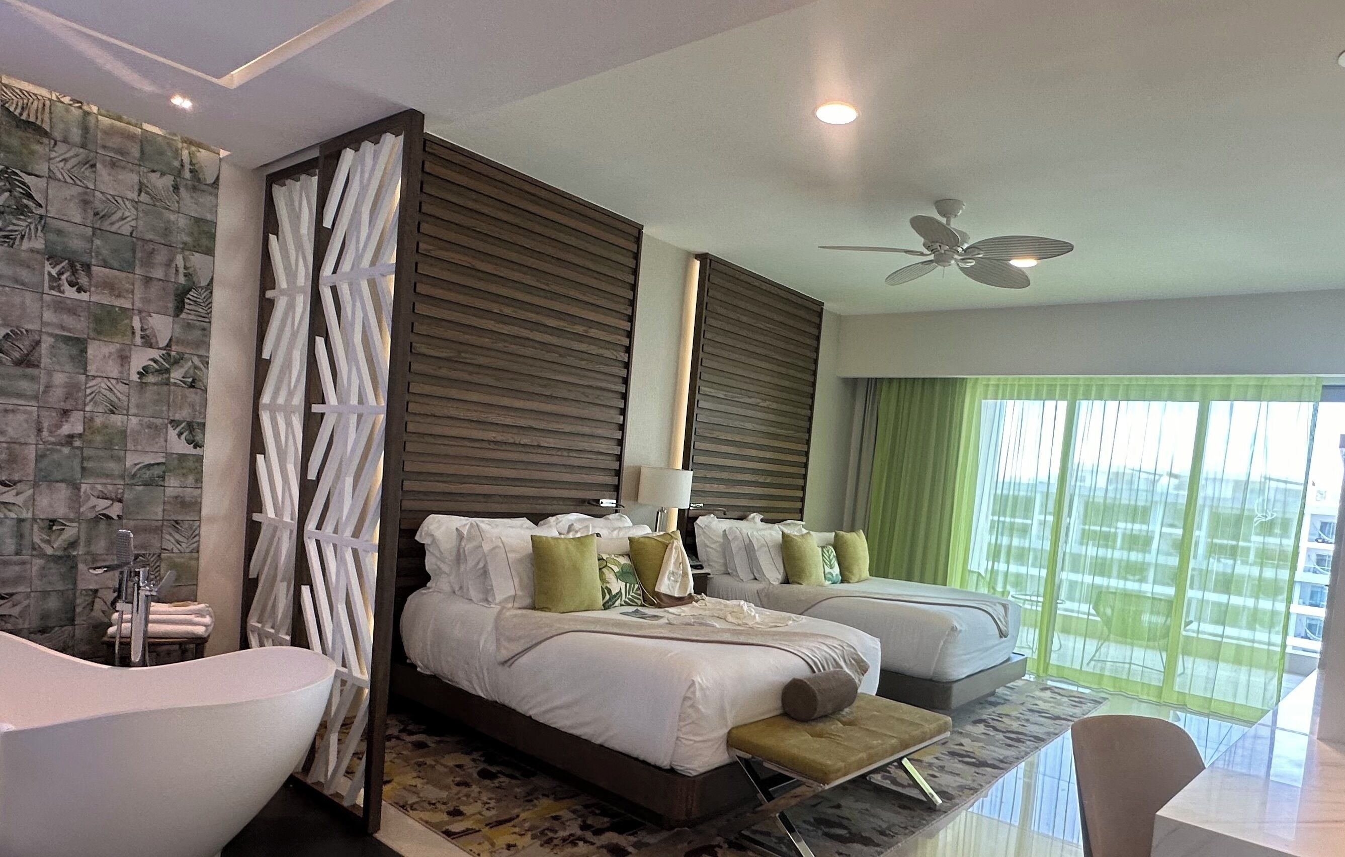 A modern hotel room with a large bed, two bedside tables, a sitting area, a bathtub, and a large window with curtains.