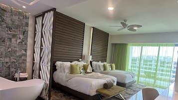 A modern hotel room with a large bed, two bedside tables, a sitting area, a bathtub, and a large window with curtains.