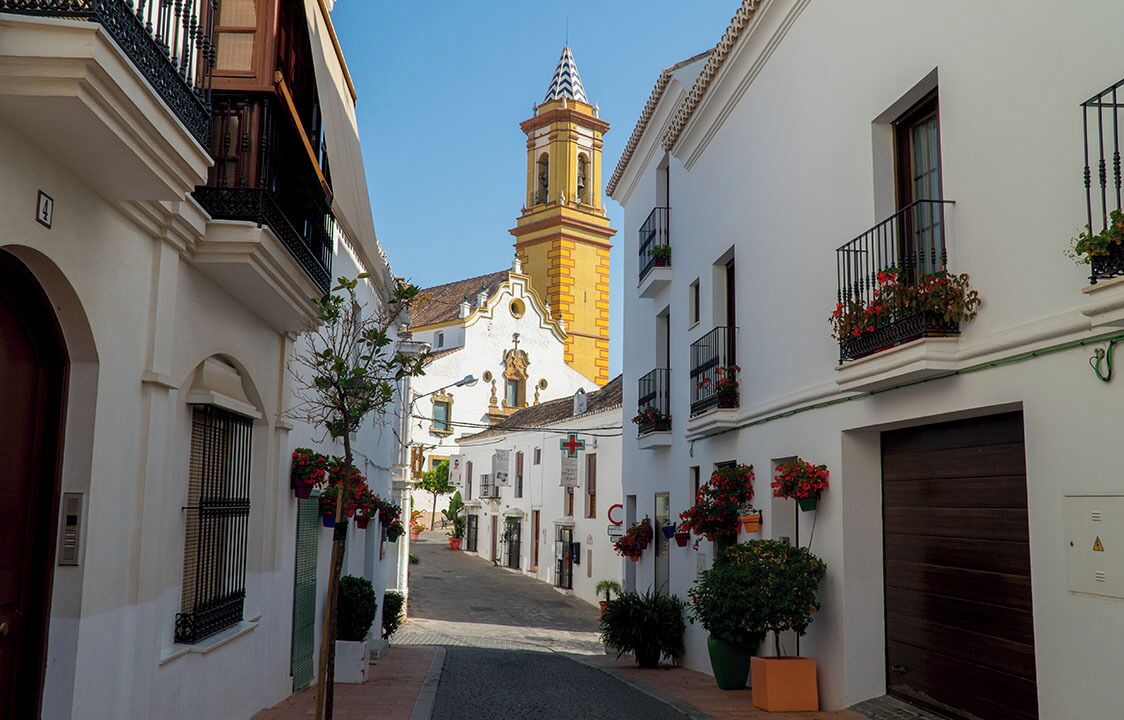 A narrow street with white buildings, a church with a bell tower, and balconies with flowers.