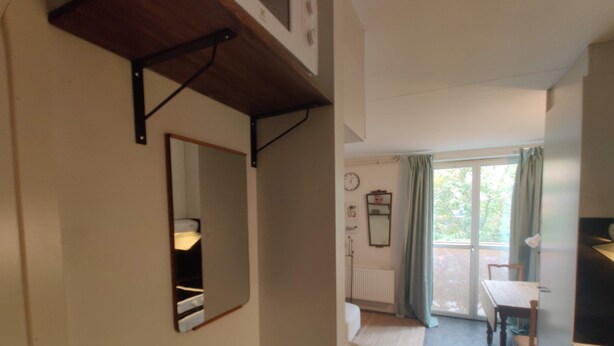 Una habitación con cama, una mesita, una silla, un radiador, una ventana con cortinas y un espejo en la pared.