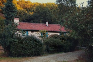 A rustic house with a red-tiled roof, surrounded by dense greenery and a gravel path.