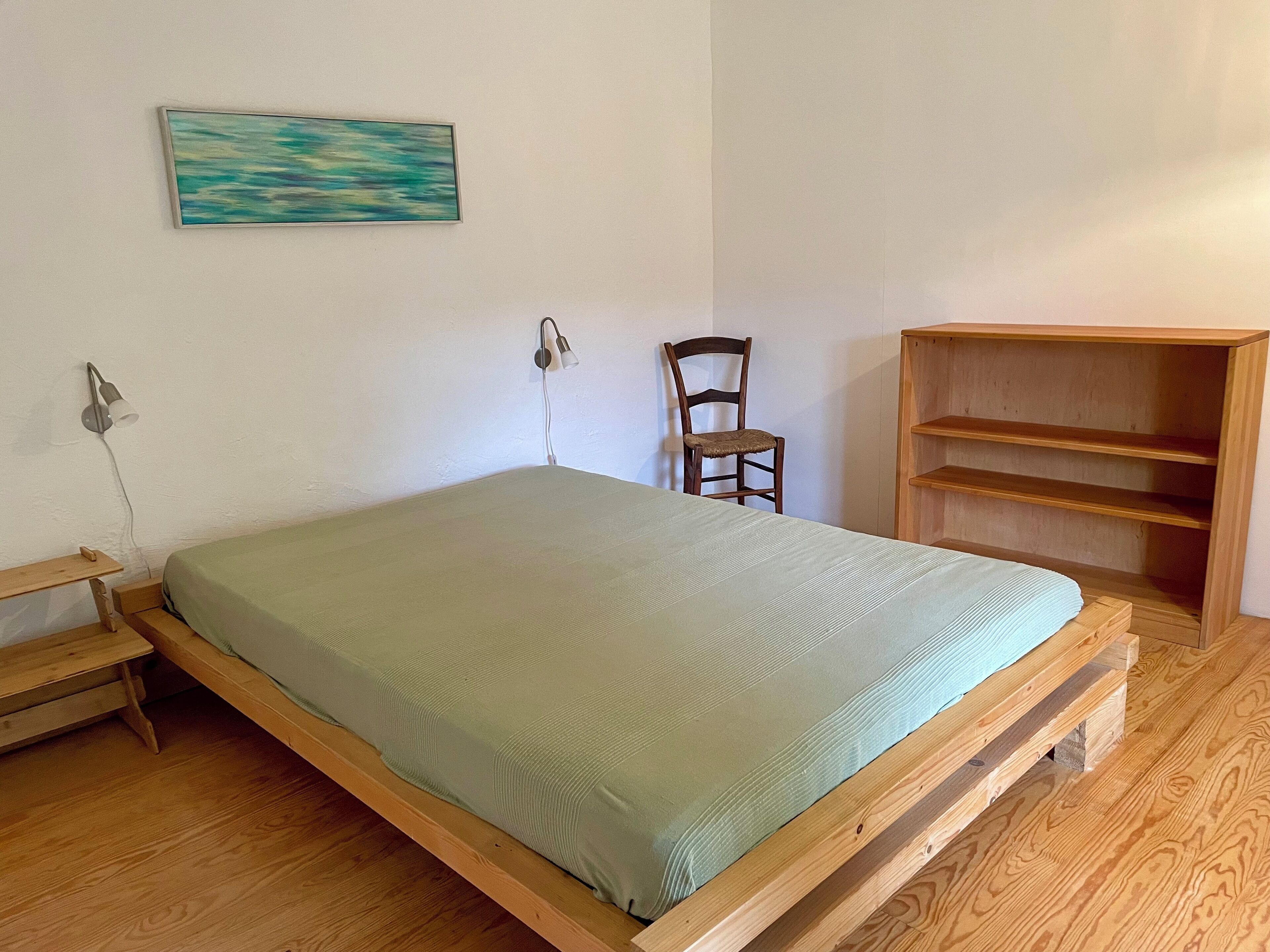 A room with a wooden bed, a chair, a wall-mounted lamp, and a wooden shelf unit.