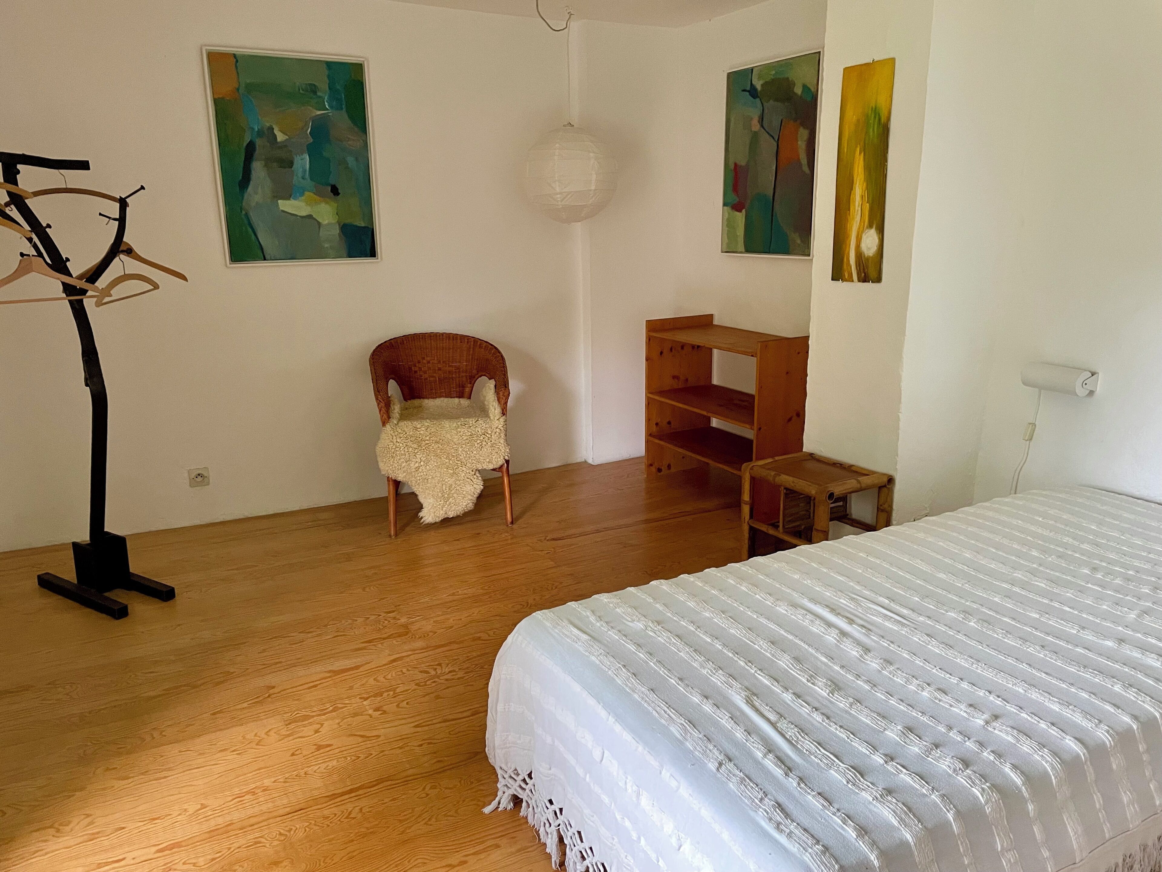 Un dormitorio con cama, una silla, estante de madera y pinturas abstractas en las paredes.