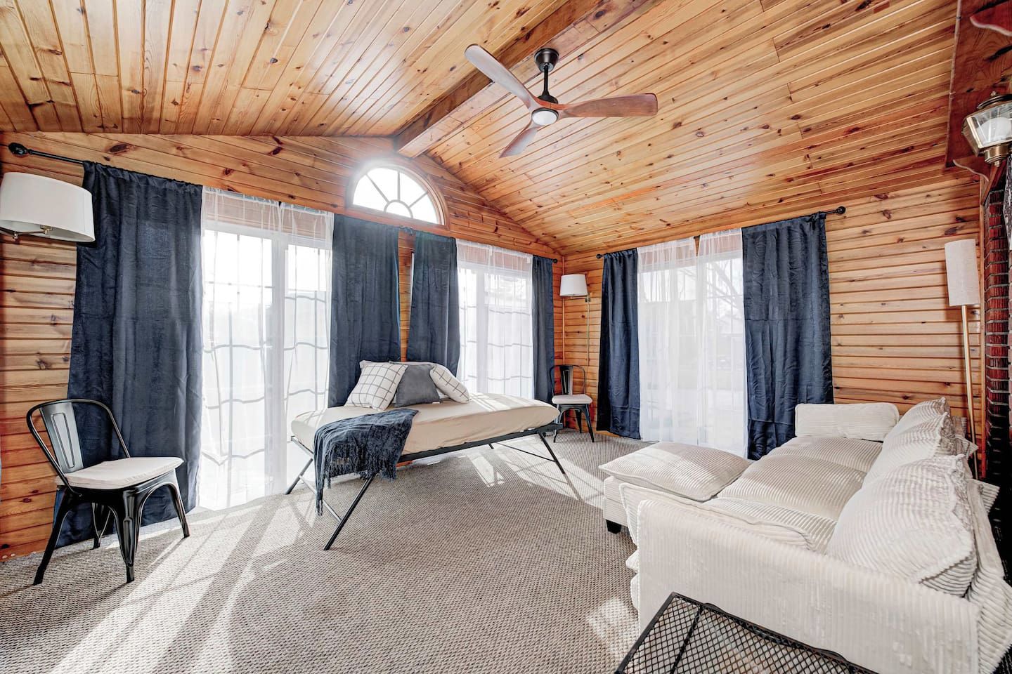 Interior de una cabaña de madera con cama, sofá y sillón, que cuenta con ventilador de techo, ventanas amplias y una estética rústica.