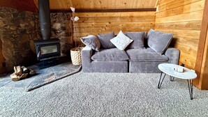 Un cuarto acogedor con pared de piedra, techo de madera, sofá gris con almohadones, una mesita blanca y chimenea con leña.
