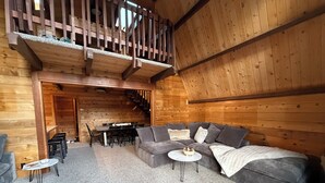 Un interior acogedor de cabaña de madera con un sofá gris, una pequeña mesa redonda y una escalera que sube a un nivel superior.