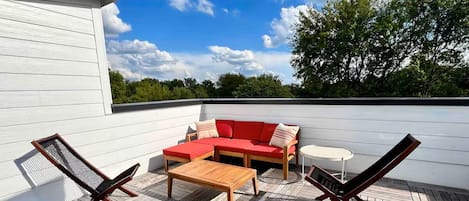 A rooftop patio with a wooden deck, a red sofa, two lounge chairs, and a small table.