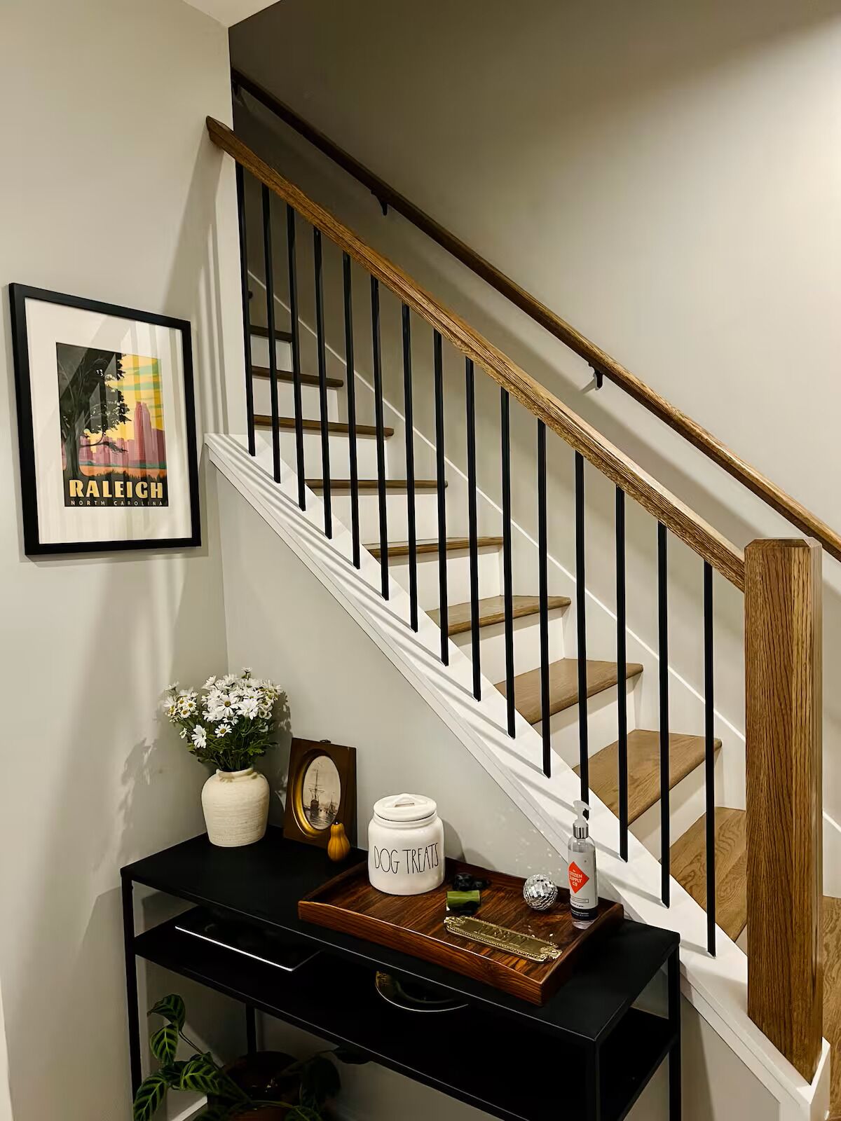 A staircase with a white railing and wooden steps, a framed picture on the wall, and a console table with a vase of flowers, a framed picture, and a container labeled "DOG TREATS".