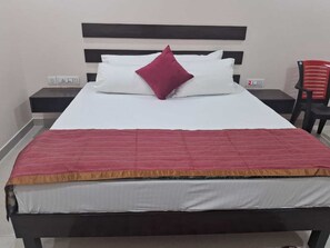 A bed with a white mattress, a red blanket, and a single red pillow. There are two bedside tables with lamps, and a red chair to the right. - Shivansh Hotel (Kotdwar)