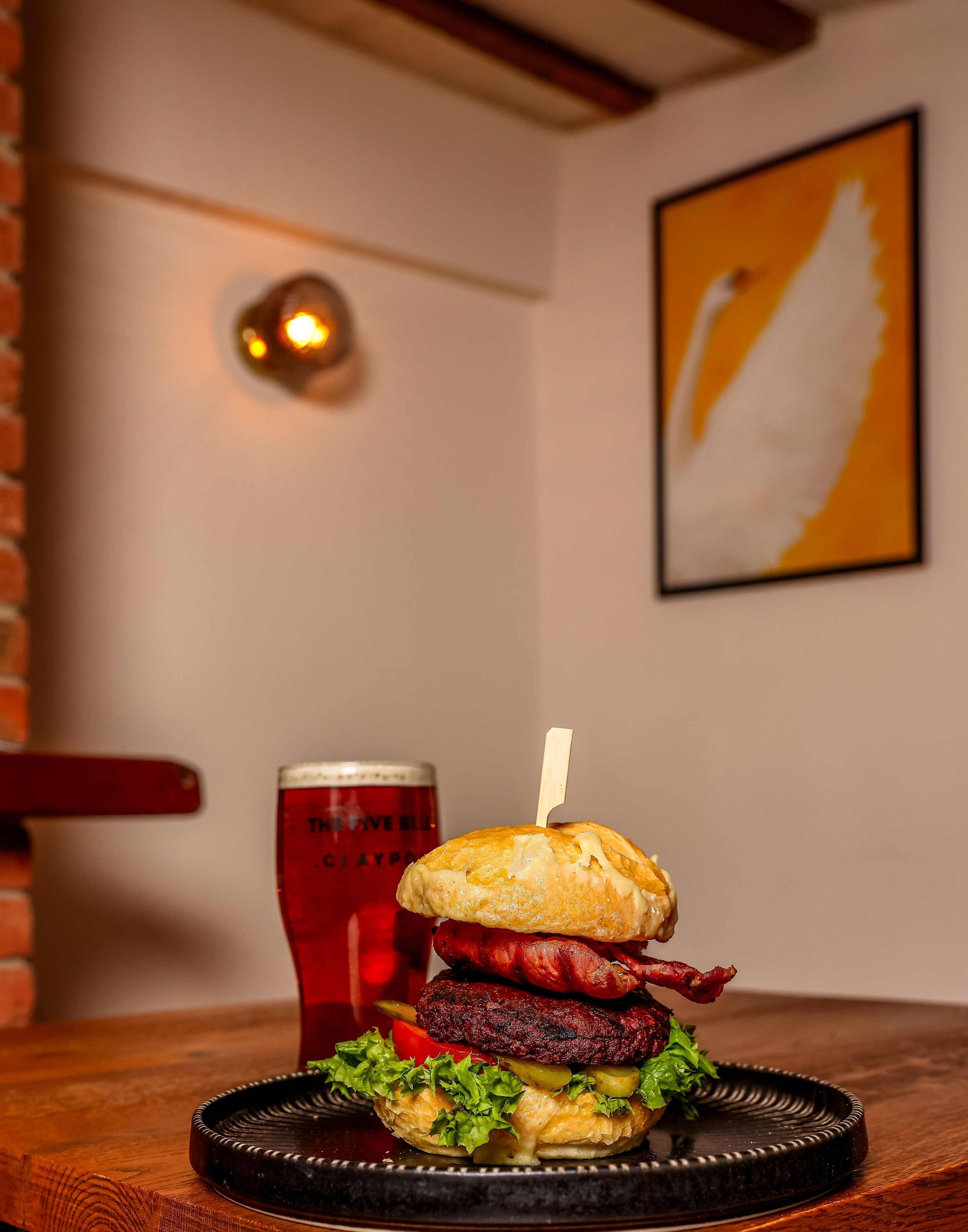 A burger with a bun, lettuce, cheese, and a sausage, served on a black plate with a glass of beer in the background.