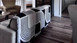 A modern living room with a sofa, armchair, and a patterned rug. There is a ceiling fan, wooden beams, and a door leading to another room. - Charming Vintage Home w/Separate Barn Guesthouse and Yard (San Antonio)